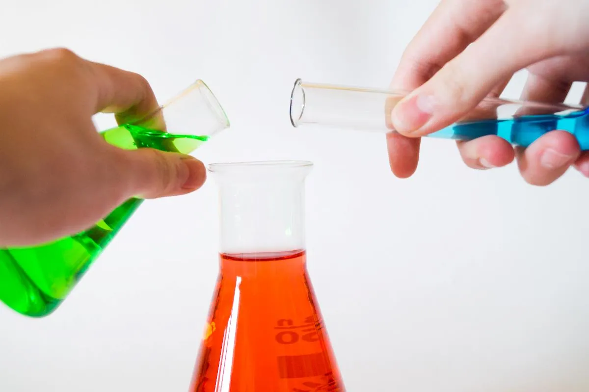 Two hands pouring coloured liquids from test tubes into a conical flask.