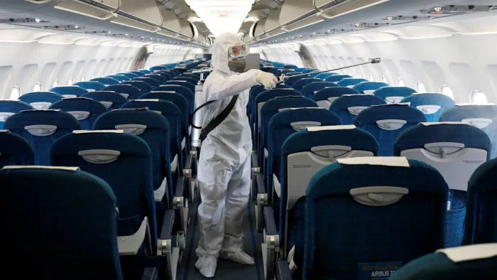 A worker in a full protective suit disinfecting an aircraft cabin between rows of seats.