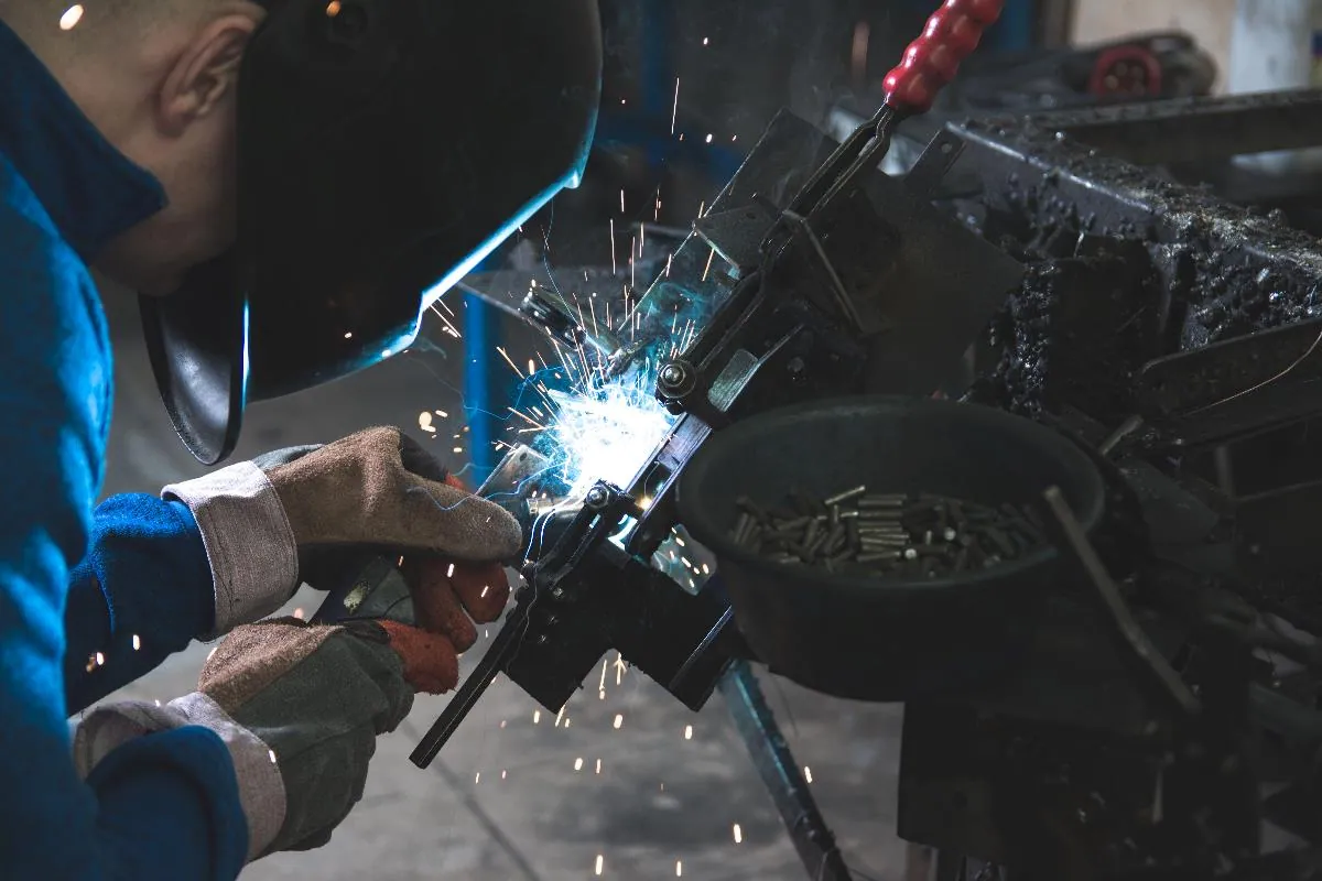 Kask ve eldiven takmış bir kaynakçı metal bir parça üzerinde çalışıyor, kıvılcımlar saçılıyor.