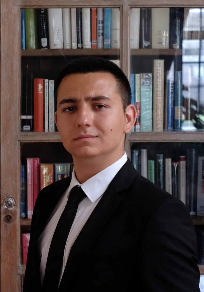 Koyu renk takım elbise ve kravatlı, bir kitaplığın önünde duran bir adamın portresi.