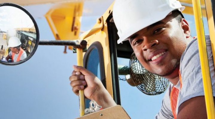 African American man, 20s, on crane at manufacturing facility.